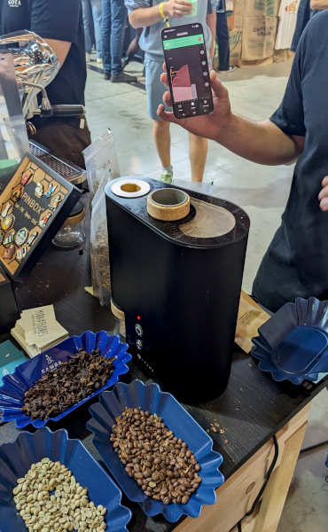 A man is holding up his smartphone showing some curve on the screen and some Ikawa machine and some roasted and fresh coffee beans are in the front